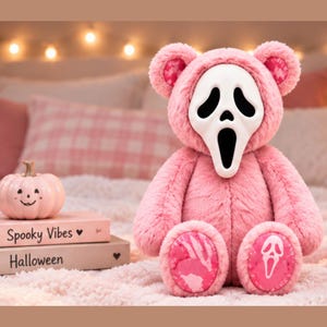 May include: A plush teddy bear with pink fur wearing a white Ghostface mask. The bear has pink patterned fabric on the inner ears and foot pads, with a white Ghostface mask icon on the right foot pad. It sits upright with a soft, fuzzy texture.