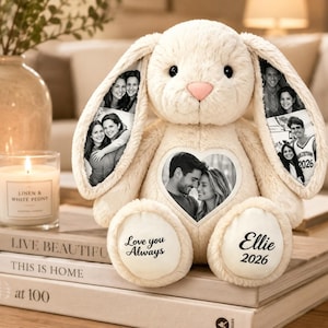 Puede incluir: Un peluche de conejo color crema con fotos en blanco y negro impresas en sus largas orejas y un parche en forma de corazón en el pecho. El pie izquierdo tiene bordado el texto Love you Always y el derecho el nombre Ellie 2026.
