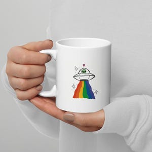 May include: White ceramic mug featuring a whimsical illustration of a UFO with a rainbow beam and a small heart. The design includes a green alien and starburst accents. The mug has a curved handle.