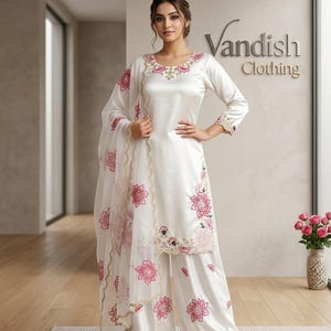 May include: A white satin outfit with pink floral embroidery, including a long-sleeved top and wide-leg trousers. A matching sheer scarf with floral detailing is draped over the shoulder. The outfit is from Vandish Clothing.
