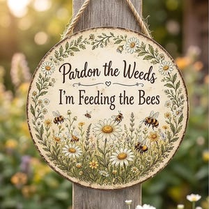 May include: A round hanging sign with a cream-colored background featuring a floral border of daisies and greenery. The text reads "Pardon the Weeds I'm Feeding the Bees" in black script and print. Several illustrated bees are depicted among the flowers.