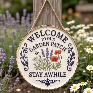 May include: A round, off-white metal sign with a distressed edge, hanging from a jute rope. It features dark blue embossed text reading "WELCOME TO OUR GARDEN PATCH" and "STAY AWHILE," with a central illustration of a bee, lavender, and poppies.
