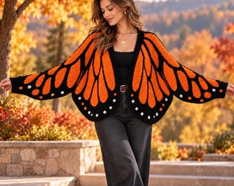May include: A black and orange butterfly wing cardigan featuring a vibrant monarch pattern with white accents along the edges, worn over a black top and dark gray jeans, creating a distinct wing-like silhouette when arms are extended.