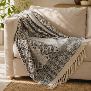 May include: A woven throw blanket featuring a black and white geometric diamond pattern, a central light blue floral stripe, and a decorative border with blue floral motifs. The blanket is finished with cream-colored fringe along the edges.