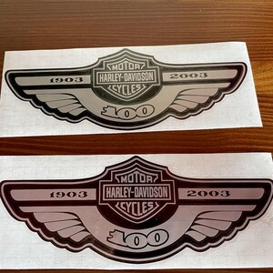 Könnte beinhalten: Zwei Aufkleber mit einem silber-schwarzen Harley-Davidson-Logo mit Flügeln. Der Text enthält "MOTOR HARLEY-DAVIDSON CYCLES" und die Jahreszahlen "1903" und "2003". Der untere Teil des Logos lautet "100".
