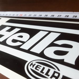 Peut inclure: Deux autocollants noirs et blancs avec le logo Hella. Les autocollants sont rectangulaires avec le mot "Hella" en grandes lettres blanches. Un mètre ruban est placé au-dessus des autocollants, indiquant des mesures jusqu'à 76 cm.