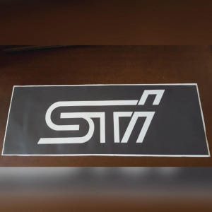 May include: A rectangular black sticker featuring the white letters "STI" in a distinctive font. The sticker has a white border and is likely for automotive use. The design is modern and clean.