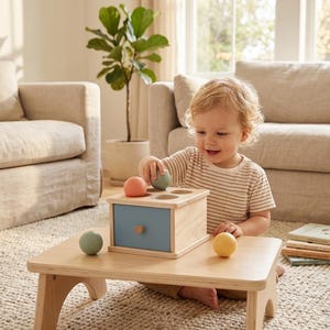 Puede incluir: Un niño juega con un juguete de madera sobre una mesa pequeña. El juguete tiene un cajón azul claro y agujeros para bolas de colores. Las bolas son rosas, verdes y amarillas. El niño lleva una camiseta a rayas.