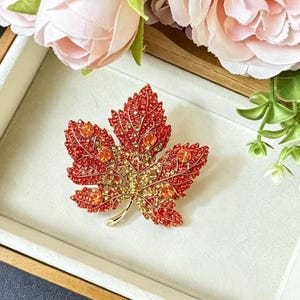 May include: A maple leaf brooch featuring a gradient of red and gold crystals. The leaf design includes five larger circular orange stones embedded within the textured, metallic surface, with a smooth gold-toned stem at the base of the pin.