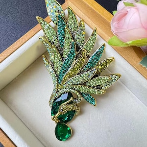 May include: A decorative gold-coloured brooch in the form of a stylized feather or wing. It is embellished with green and yellow rhinestones, and features a large emerald-green, teardrop-shaped stone. The brooch is displayed in a white-lined box.