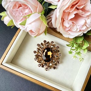 May include: A gold-toned brooch with a large, oval, dark brown center stone, surrounded by smaller, lighter brown stones in a floral design. The brooch is set on a white surface inside a wooden box, with pink roses in the background.