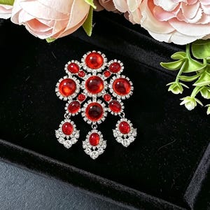 May include: A silver-toned brooch featuring a cluster of round, deep red cabochon stones surrounded by small, clear sparkling crystals. The design includes three dangling elements at the bottom, each set with a single red stone and clear crystals.