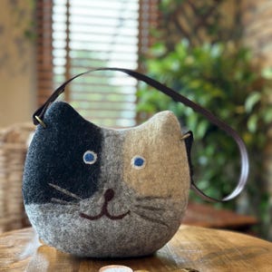 May include: A gray, black, and cream-colored cat-shaped handbag with a brown strap. The bag has blue eyes and a stitched smile. A gold wristwatch with a white face rests on a wooden surface.