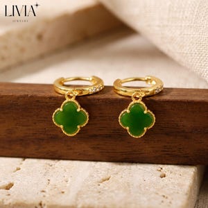 May include: A pair of gold hoop earrings featuring a dangling green clover-shaped charm. Each hoop is accented with small clear stones along the outer edge. The jewellery is branded with the text LIVIA JEWELRY in the upper left corner.