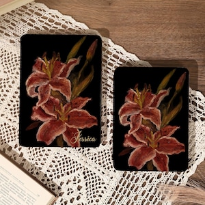 May include: Two black rectangular objects with a floral design. The design features red and orange lilies with green stems and leaves. The name "Jessica" is written in cursive on the larger object. The objects are on a white lace surface.