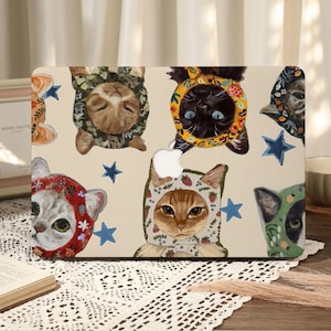 Könnte beinhalten: Eine Laptop-Hülle mit einem skurrilen Design von Katzen, die verschiedene gemusterte Hüte tragen. Die Katzen sind in verschiedenen Farben dargestellt, mit blauen Sternakzenten auf cremefarbenem Hintergrund. Das Apple-Logo ist sichtbar.