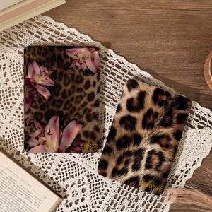 Könnte beinhalten: Zwei rechteckige E-Reader-Hüllen mit Tiermustern. Eine Hülle zeigt ein braunes Leopardenmuster mit rosa Lilien und dem Namen Lizzie in goldener Schrift. Die andere Hülle hat ein braun-schwarzes Leopardenmuster mit dem Namen Deborah.