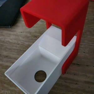 May include: A red and white 3D-printed object with a rectangular top and a sloped, open-ended tray. The tray has a circular hole near the bottom. The object appears to be a desk organizer or a similar item.