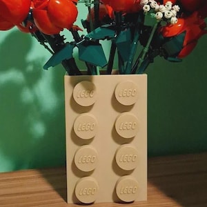 Könnte beinhalten: Eine rechteckige beige Vase aus Kunststoff-Bausteinen mit dem eingeprägten Wort LEGO auf jedem der acht kreisförmigen Noppen. Die Vase enthält einen Strauß aus roten Kunststoffrosen mit grünen Stielen und Blättern sowie kleinen weißen Blüten.