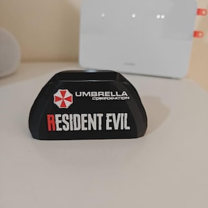 May include: A black, curved, semi-circular stand featuring the red and white Umbrella Corporation logo on the left side. The text "UMBRELLA CORPORATION" is printed in white, and the words "RESIDENT EVIL" are displayed in large white and red letters.