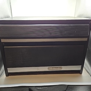 Pode incluir: Uma unidade de armazenamento Nintendo retangular preta com duas gavetas horizontais, painéis frontais estriados e acabamentos prateados. A gaveta inferior exibe o logótipo da Nintendo num emblema oval prateado na faixa horizontal inferior.