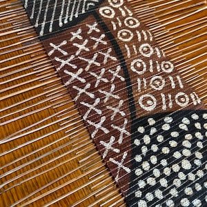 May include: A close-up of a brown and black fabric with white geometric patterns. The fabric is woven on a loom with thin wooden rods.