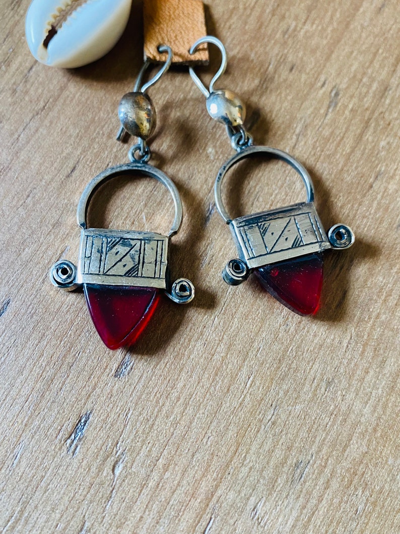 May include: A pair of silver-toned earrings with a geometric design. Each earring features a rectangular top with etched lines and a teardrop-shaped, deep red glass pendant. The earrings have a hook closure and a small, round bead at the top.