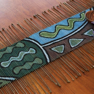 May include: A woven tapestry with a geometric pattern in shades of blue, green, brown, and white. The tapestry has a fringe on one side and is lying on a wooden surface.