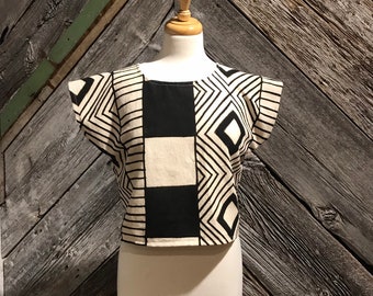 May include: A black and white geometric patterned cropped top with short sleeves. The top is on a white mannequin against a wood background.