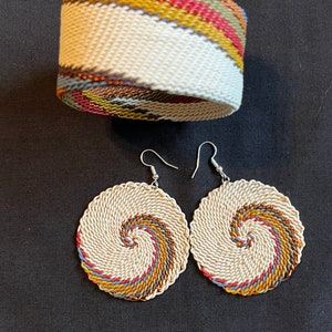 May include: A set of earrings and a bracelet made of woven white cord with colorful stripes. The earrings are circular and have a spiral design. The bracelet is a wide band with a diagonal stripe pattern.