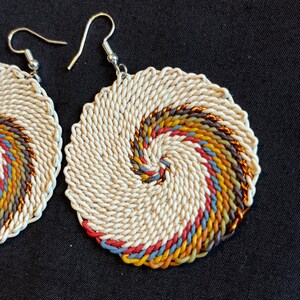 May include: A pair of white woven earrings with a colorful spiral design. The earrings are made of a white cord with a spiral of brown, orange, yellow, blue, and red cord.