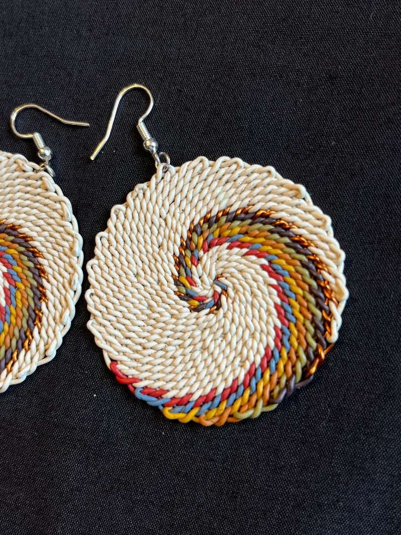 May include: A pair of white woven earrings with a colorful spiral design. The earrings are made of a white cord with a spiral of brown, orange, yellow, blue, and red cord.