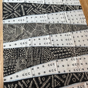May include: A white fabric with a repeating pattern of black geometric shapes and lines. The design includes diagonal stripes filled with various patterns, such as dots, dashes, and curved lines. The fabric has a textured appearance.