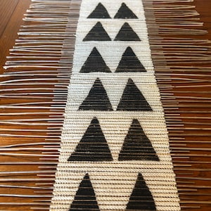 May include: A white table runner with black triangle designs. The runner has fringe on the edges.