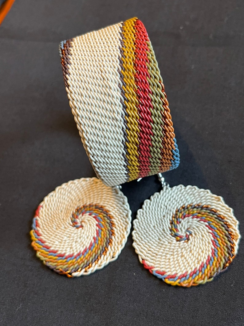 May include: A set of white woven earrings and a bracelet with a multi-colored striped design. The earrings are round and have a spiral pattern. The bracelet is wide and has a straight design.