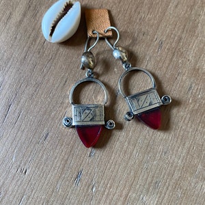 May include: Silver-toned earrings with a red, triangular gem and a decorative silver top. The earrings hang from a small leather square. A white seashell is next to the earrings. The earrings are on a wooden surface.