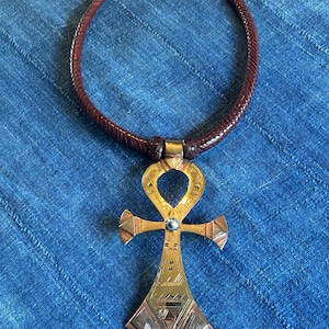 May include: A brown leather necklace with a gold-colored Ankh pendant. The pendant has intricate geometric designs and a small silver-colored bead. The necklace is displayed on a blue fabric background.