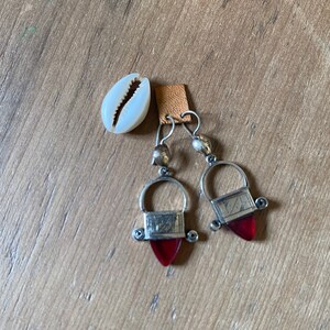 May include: Silver-toned earrings with red teardrop-shaped pendants and a small, white seashell. The earrings have a unique design with a rectangular top and a curved handle. The seashell is placed on a wooden surface.