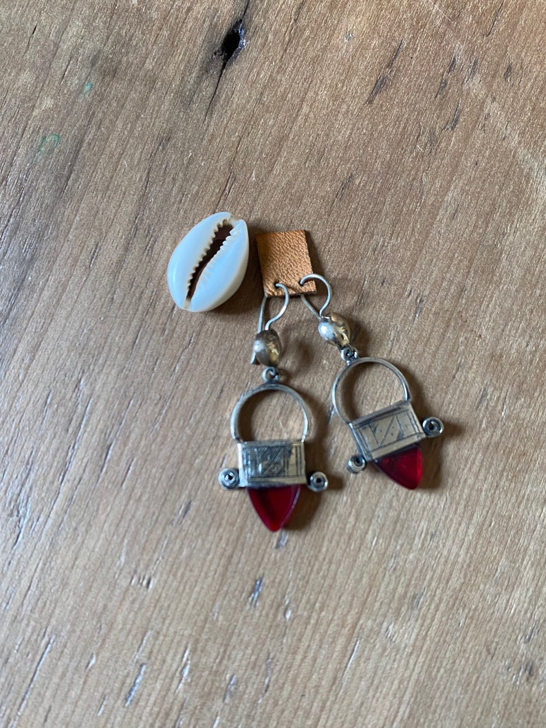 May include: Silver-toned earrings with red teardrop-shaped pendants and a small, white seashell. The earrings have a unique design with a rectangular top and a curved handle. The seashell is placed on a wooden surface.