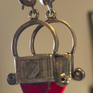 May include: Silver-toned dangle earrings with a unique design. Each earring features a small, round top, a circular hoop, and a rectangular element with a red, heart-shaped pendant. The earrings have an antique or vintage aesthetic.