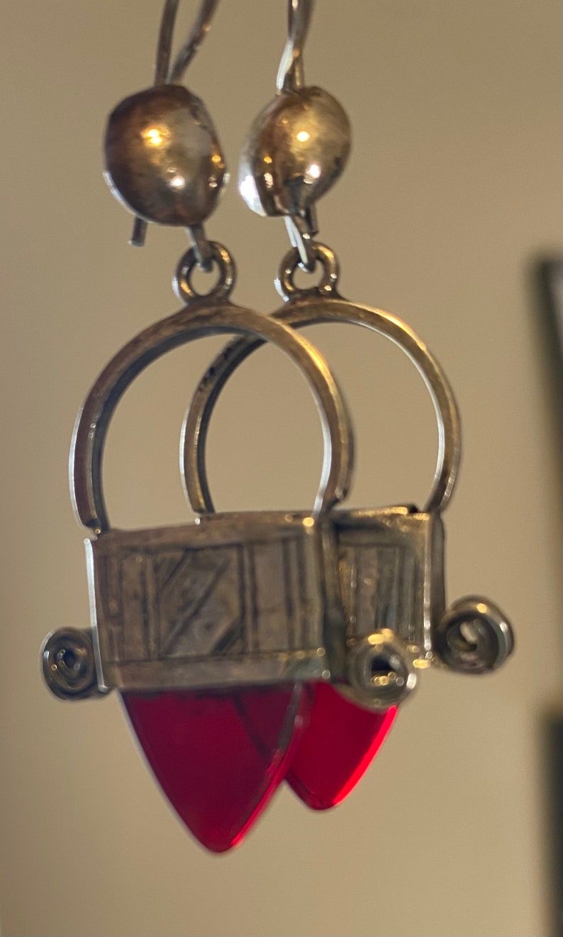 May include: Silver-toned dangle earrings with a unique design. Each earring features a small, round top, a circular hoop, and a rectangular element with a red, heart-shaped pendant. The earrings have an antique or vintage aesthetic.