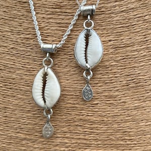 May include: Two silver necklaces with cowrie shell pendants and small silver charms. The necklaces are on a brown background.
