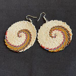 May include: A pair of circular earrings with a woven design. The earrings are made of white and brown string with a spiral pattern. The earrings have silver hooks.