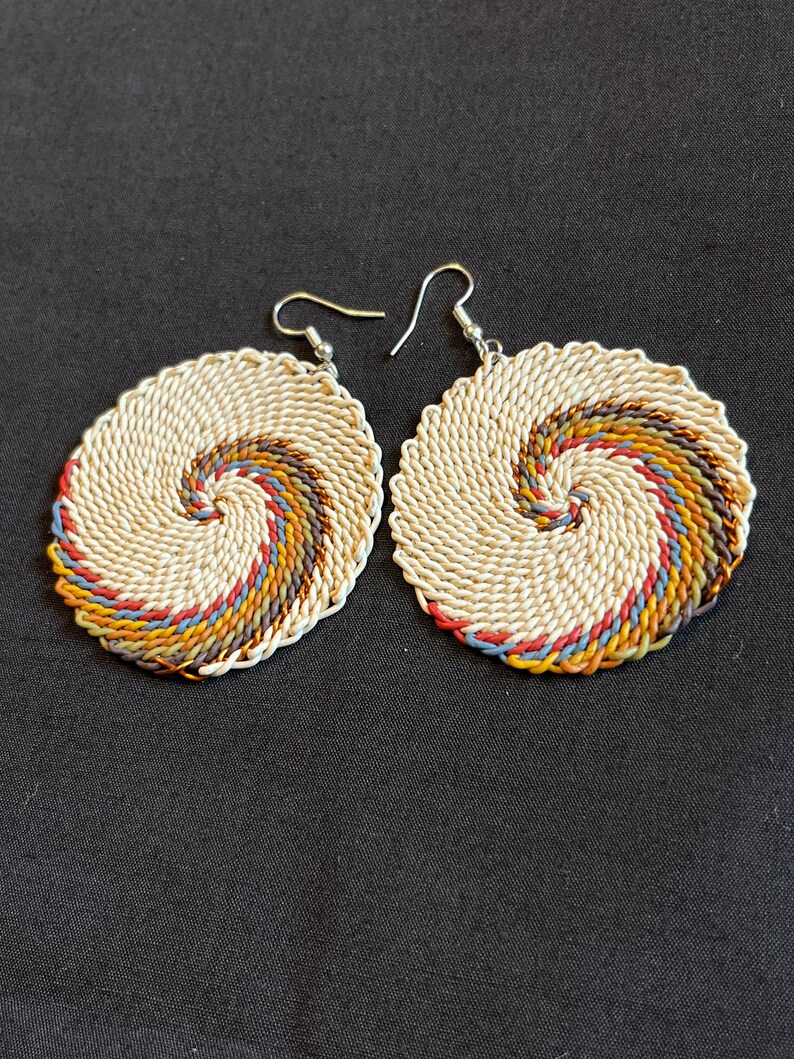 May include: A pair of circular earrings with a woven design. The earrings are made of white and brown string with a spiral pattern. The earrings have silver hooks.