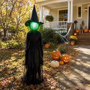 May include: A tall standing witch decoration featuring a black pointed hat and a long black dress with a sheer overlay. The head is a glowing green orb. The figure is positioned outdoors on a lawn covered in fallen autumn leaves during the daytime.
