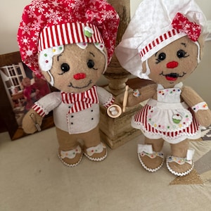May include: Two gingerbread-style plush dolls dressed as chefs. The left doll wears a red snowflake-patterned hat and striped necktie. The right doll wears a white checkered hat and apron with a cupcake motif. Both have embroidered faces.