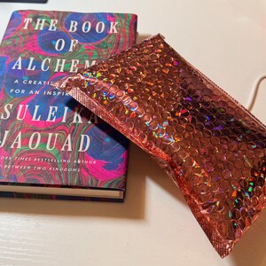 May include: A hardback book titled The Book of Alchemy by Suleika Jaouad with a colourful marbled cover featuring swirls of blue, pink, and green, positioned next to a rectangular metallic rose gold bubble mailer with a holographic finish.