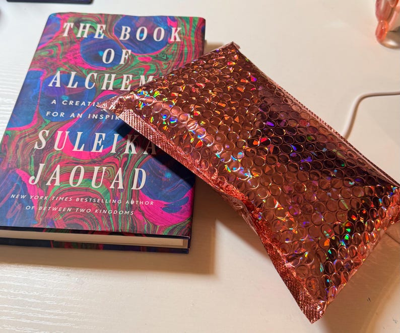 May include: A hardback book titled The Book of Alchemy by Suleika Jaouad with a colourful marbled cover featuring swirls of blue, pink, and green, positioned next to a rectangular metallic rose gold bubble mailer with a holographic finish.
