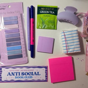 May include: A collection of stationery items including a pack of Mr. Pen sticky index tabs, a pink pen, a blue pen, a purple marker, a pink and blue dual-tip marker, a Bigelow green tea bag, a lavender hair claw, a small pink pouch with washi tape, a lined notepad, a square purple sticky note, a square hot pink sticky note, a tube of Sakura Garden hand cream, and a rectangular bookmark with the text "Lifetime Membership Anti Social Book Club."