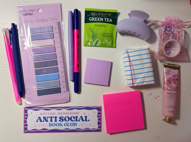 May include: A collection of stationery items including a pack of Mr. Pen sticky index tabs, a pink pen, a blue pen, a purple marker, a pink and blue dual-tip marker, a Bigelow green tea bag, a lavender hair claw, a small pink pouch with washi tape, a lined notepad, a square purple sticky note, a square hot pink sticky note, a tube of Sakura Garden hand cream, and a rectangular bookmark with the text "Lifetime Membership Anti Social Book Club."